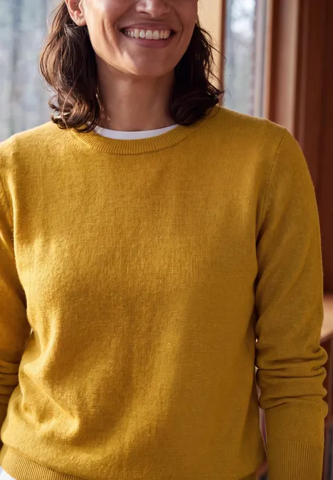 Eine lächelnde Frau trägt einen gelben Pullover in einem gemütlichen Innenraum.
