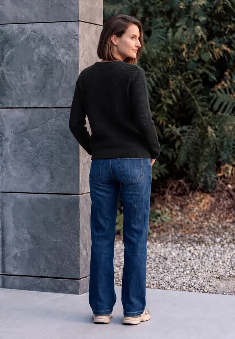 Pullover mit Rundhals und Struktur Black Frau im schwarzen Pullover und blauen Jeans lehnt an grauer Wand, blickt auf Kies und Büsche im Hintergrund.