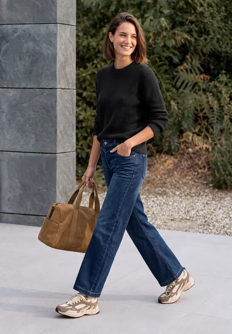 Pullover mit Rundhals und Struktur Black Eine junge Frau in schwarzem Pullover, blauen Jeans und Sneakers geht lächelnd mit einer Tasche spazieren.