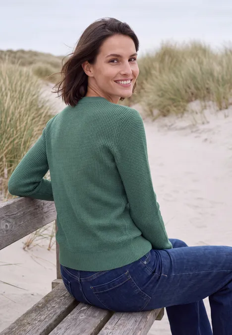 Pullover mit Rundhals und Struktur jewel green melange Frau sitzt auf einer Strandbank vor Dünengras, blickt zurück, trägt grünen Pullover und Jeans.