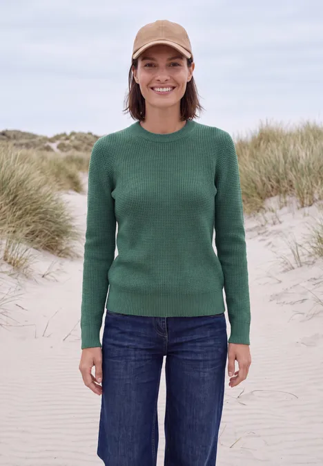 Pullover mit Rundhals und Struktur jewel green melange Eine Frau trägt grünen Pullover, Jeans und beige Mütze, lächelt vor Dünen am Strand.