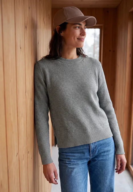 Pullover mit Rundhals und Struktur sporty grey melange Eine Frau lehnt in grauem Pullover, blauer Jeans und Cap an einer Holzverkleidung und lächelt.