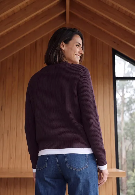 Pull à col rond et texturé eggplant red melange Femme en pull violet et jean regardant par-dessus l’épaule dans une maison en bois.