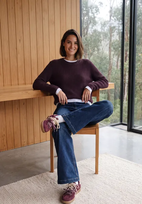 Pull à col rond et texturé eggplant red melange Jeune femme assise détendue sur une chaise en bois, devant un mur clair, vêtue d'un pull violet, jean et baskets.