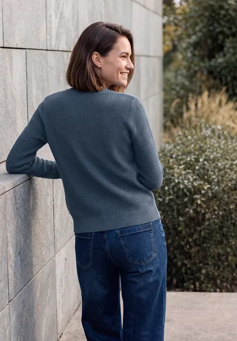Eine lächelnde Frau in grauem Pullover und blauer Jeans lehnt sich an eine Steinwand und schaut zur Seite.