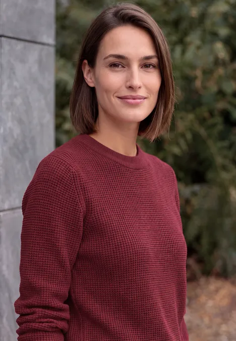 Eine Frau mit kinnlangen braunen Haaren und bordeauxrotem Pullover im Freien.