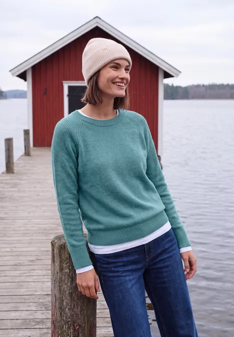Pull à col rond et mélange de textures multi lapis melange Femme souriante en pull vert, jean et bonnet, debout sur un ponton devant un abri à bateaux rouge au bord du lac.