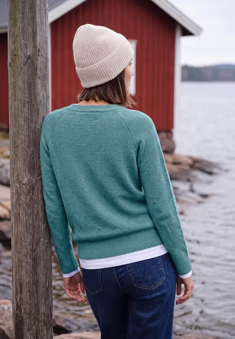 Pull à col rond et mélange de textures multi lapis melange Personne en pull vert et bonnet, vue de dos, près de l'eau devant une maison rouge.