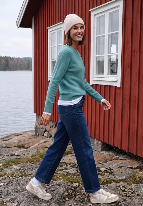 Pull à col rond et mélange de textures multi lapis melange Une femme en jean et pull sourit en marchant près d'une maison rouge au bord du lac.