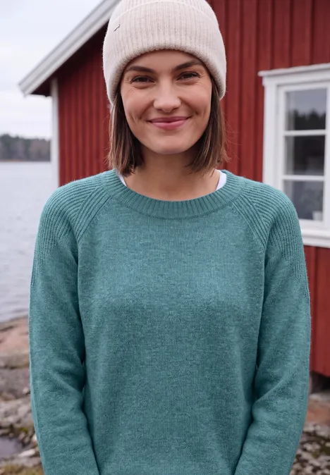 Pull à col rond et mélange de textures multi lapis melange Une femme souriante en bonnet et pull devant une maison rouge au bord de l'eau.
