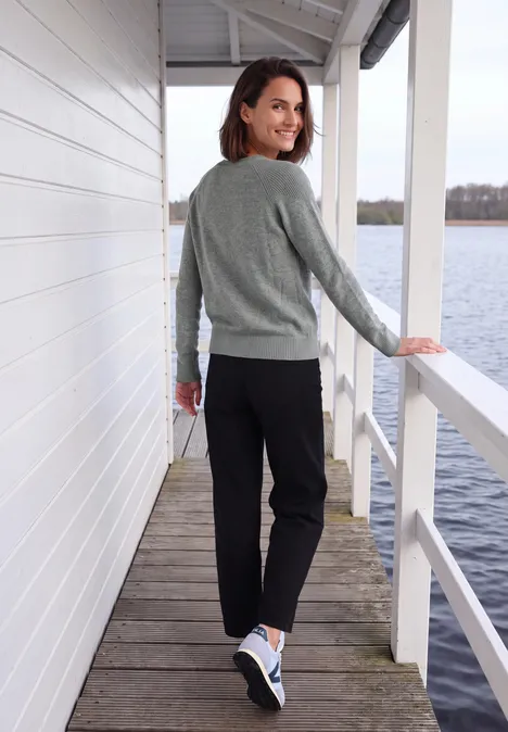 Een vrouw in een grijze trui en zwarte broek loopt over een steiger bij het water en kijkt glimlachend achterom.