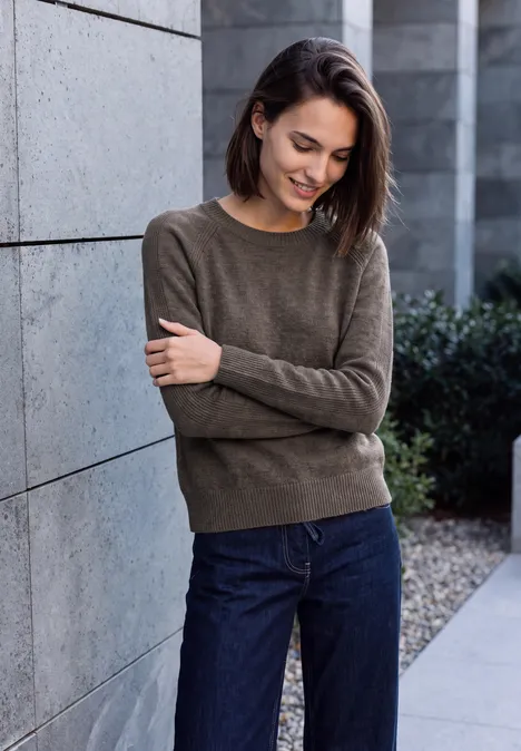 Pullover mit Rundhals und Strukturmix mushroom taupe melange Junge Frau mit braunen Haaren lehnt lächelnd an Betonwand, trägt Pulli und Jeans.