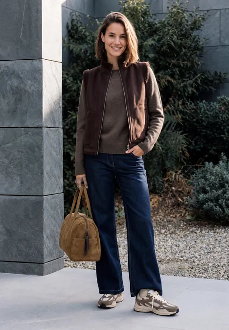 Pullover mit Rundhals und Strukturmix mushroom taupe melange Frau in brauner Jacke, dunkelblauen Jeans und Sneakers mit handlicher Tasche vor modernem Gebäude.