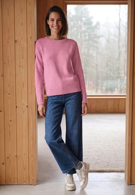Een lachende vrouw in een roze trui, wijde jeans en witte sneakers staat in een lichte houten ruimte.