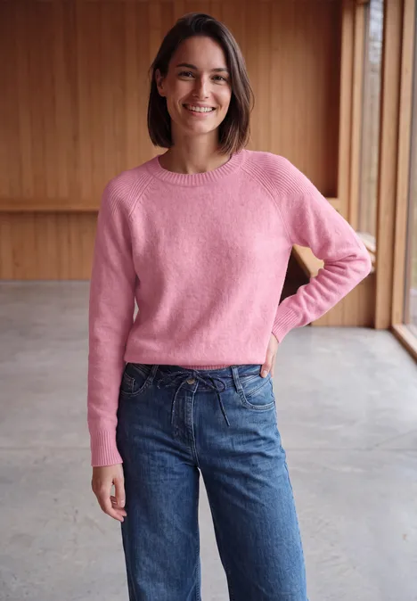 Een lachende vrouw in een roze trui en spijkerbroek staat voor een houten wand.