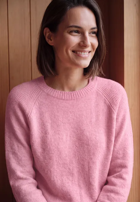 Een glimlachende vrouw in een roze trui zit voor een houten wand.