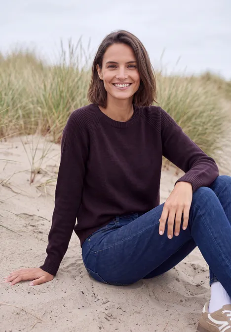 Eine lächelnde Frau sitzt entspannt im Sand vor Dünengras, trägt einen dunklen Pullover und blaue Jeans.