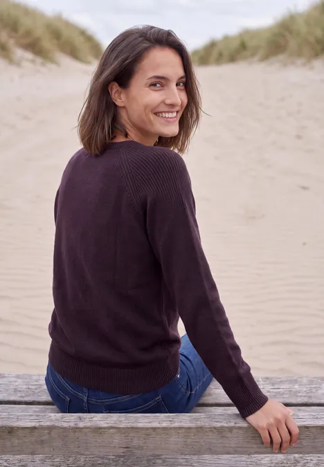 Eine Frau sitzt auf einer Holzbank am Strand, schaut lächelnd über die Schulter zu den Dünen.