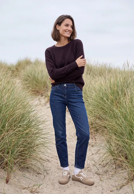 Eine Frau in Jeans und Pullover geht lächelnd zwischen Dünen am Strand spazieren.
