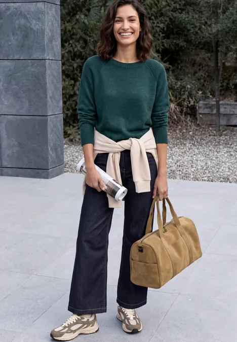 Frau mit grünem Pullover und Jeans steht vor einem Gebäude, trägt Sneakers und hält Tasche und Beutel.