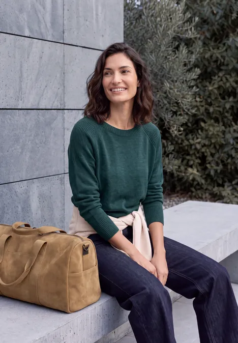 Junge Frau in grünem Pullover und Jeans sitzt vor einer Mauer, lächelnd neben einer braunen Reisetasche.
