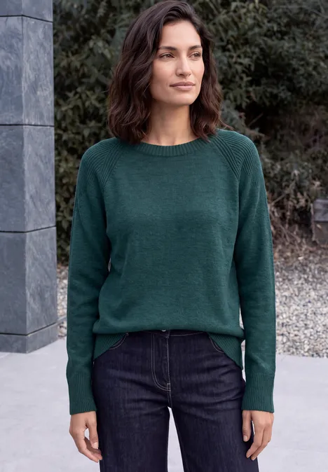 Frau in grünem Pullover und Jeans steht vor Mauer und Büschen im Freien.