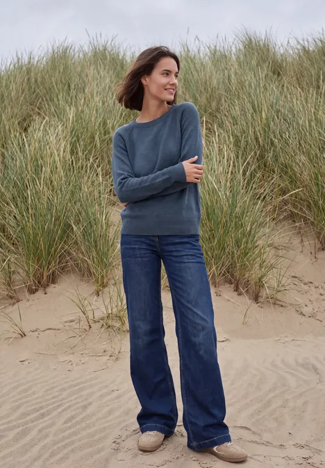 Frau steht am Strand vor Dünen, trägt blauen Pullover und Jeans, lächelnd im Wind.