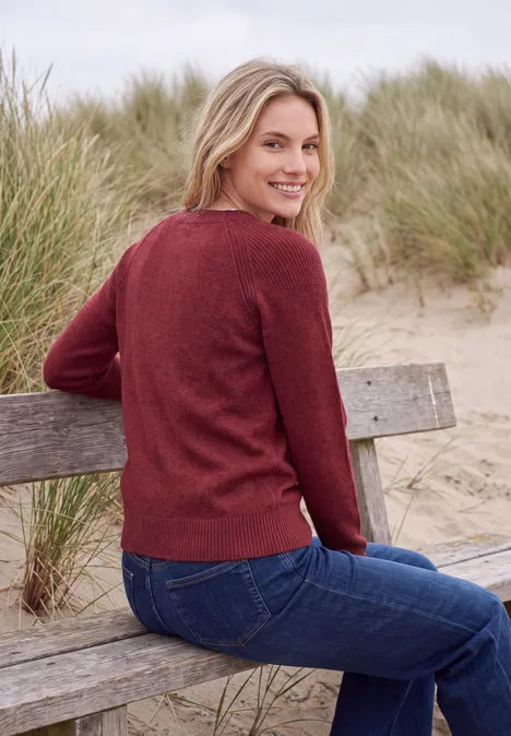 Pullover mit Rundhals und Strukturmix rhubarb melange Frau mit rotem Pullover und Jeans sitzt auf einer Holzbank in den Dünen und lächelt.