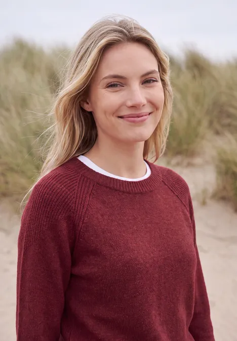 Pullover mit Rundhals und Strukturmix rhubarb melange Eine lächelnde Frau in rotem Pullover steht vor grasbedeckten Dünen am Meer.