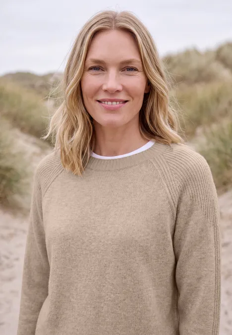 Pull à col rond et mélange de textures cashew beige melange Une femme blonde en pull beige sourit devant un paysage de dunes.