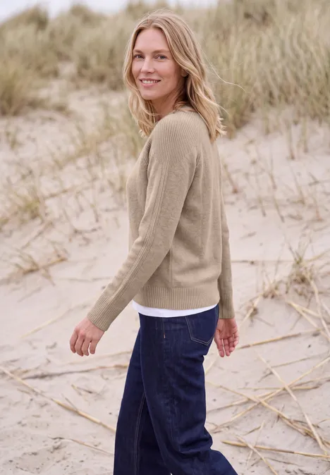 Pull à col rond et mélange de textures cashew beige melange Une femme souriante en pull beige et jean foncé se promène sur la plage parmi les dunes.