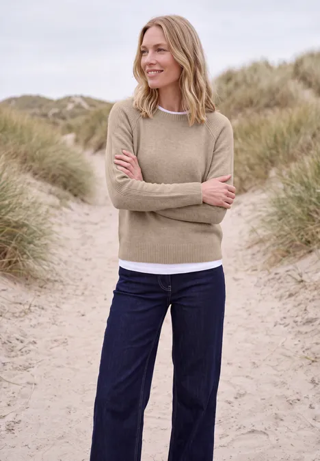 Pull à col rond et mélange de textures cashew beige melange Une femme souriante en pull beige et jean bleu, bras croisés, debout sur des dunes de sable avec des herbes.