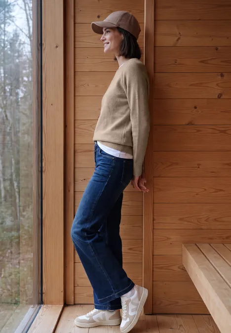 Pull à col rond et mélange de textures cashew beige melange Vue latérale d’une femme en jean, pull beige et casquette, souriant, appuyée près d’une fenêtre dans un intérieur en bois.