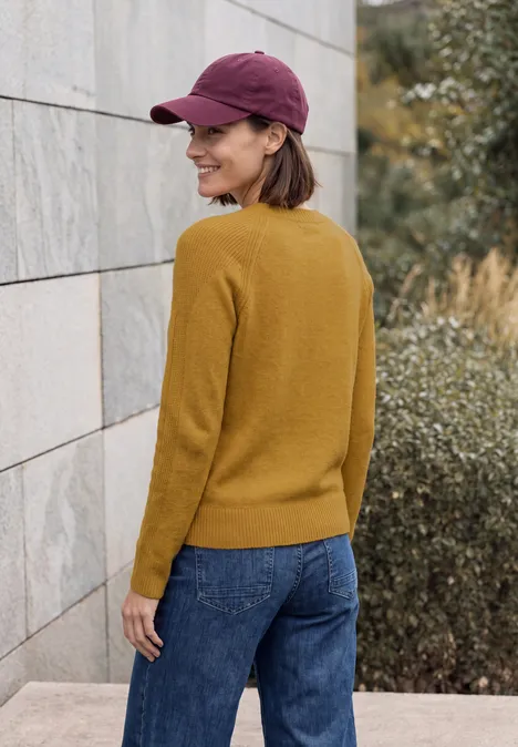 Pull col rond à textures contrastées golden honey melange Femme en pull jaune, jean et casquette violette, souriante, regardant par-dessus l'épaule devant un mur et des buissons.