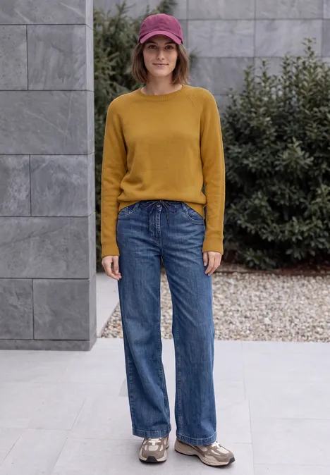 Pull col rond à textures contrastées golden honey melange Une femme portant un pull moutarde, un jean large, une casquette et des baskets devant un bâtiment moderne.