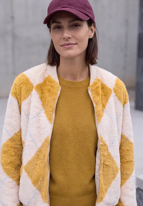 Pull col rond à textures contrastées golden honey melange Une femme porte une veste à losanges jaune et blanche, un pull moutarde et une casquette rose.