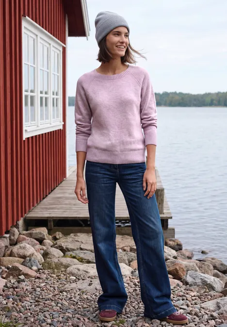Vrouw in roze trui en muts, met een glimlach, in jeans voor een rood huisje aan het meer.