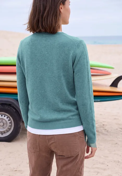 Langarm Pullover mit V-Neck multi lapis melange Frau in grünem Pullover und brauner Hose am Strand vor bunten Surfboards.