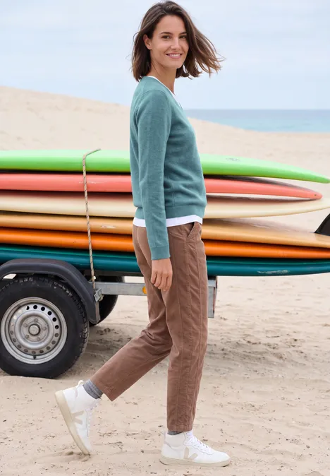 Langarm Pullover mit V-Neck multi lapis melange Eine Frau steht am Strand neben bunten Surfbrettern, trägt lässige Kleidung und lächelt.