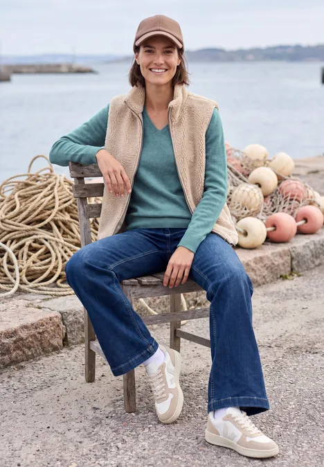 Langarm Pullover mit V-Neck multi lapis melange Junge Frau mit Cap, Fleece-Weste und Jeans sitzt lächelnd an einem Hafen mit Bojen und Seilen.