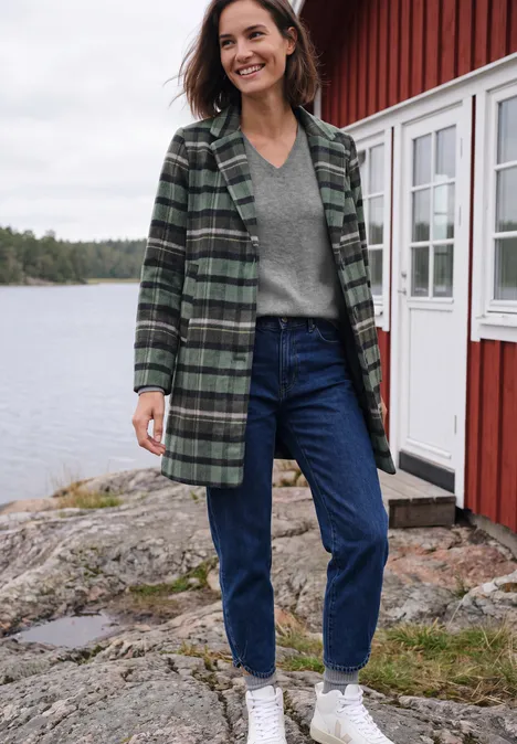 Langarm Pullover mit V-Neck sporty grey melange Eine Frau steht lächelnd vor einem roten Holzhaus am See, trägt Jeans, grauen Pullover und karierten Mantel.