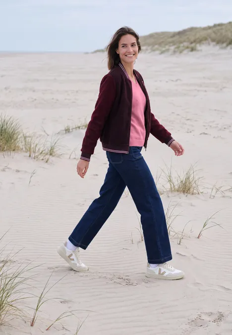Langarm Pullover mit V-Neck dark blossom melange Frau in roter Jacke und Jeans spaziert lächelnd über Dünen am Strand.