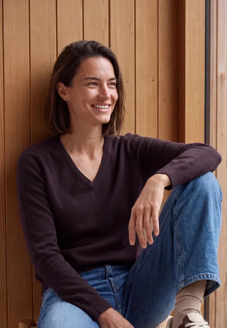 Langarm Pullover mit V-Neck eggplant red melange Eine lächelnde Frau sitzt entspannt vor einer Holzverkleidung, trägt Pullover und Jeans.