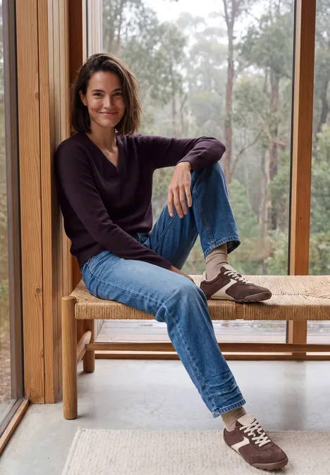 Langarm Pullover mit V-Neck eggplant red melange Frau in lässigem Outfit sitzt auf Holzbank vor großem Fenster mit Waldsicht.