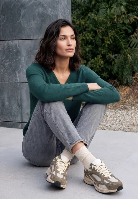 Langarm Pullover mit V-Neck dark sea green melange Frau in grüner Jacke und Jeans sitzt entspannt auf dem Boden, lehnt an einer Wand.
