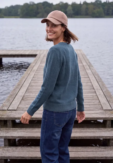 Eine lächelnde Frau mit Mütze und Pullover steht auf einem Steg am ruhigen See.