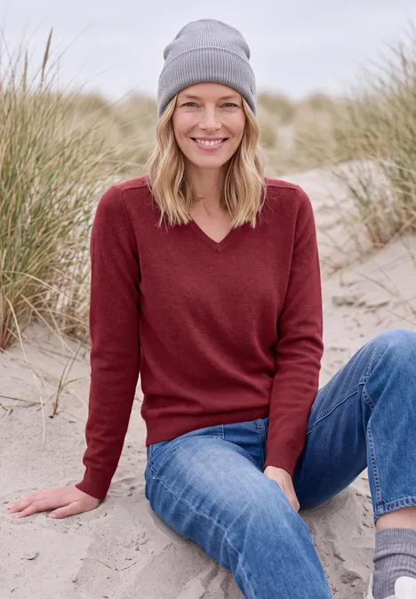 Langarm Pullover mit V-Neck rhubarb melange Eine lächelnde Frau in Mütze, rotem Pullover und Jeans sitzt im Sand vor Dünen.