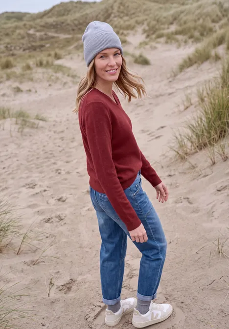 Langarm Pullover mit V-Neck rhubarb melange Junge Frau in rotem Pullover, Jeans, grauer Mütze steht lachend in Dünen.