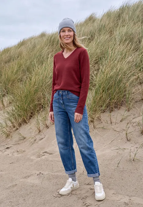 Langarm Pullover mit V-Neck rhubarb melange Eine lächelnde Frau mit grauer Mütze, rotem Pullover, blauen Jeans und weißen Schuhen steht auf einer Sanddüne.