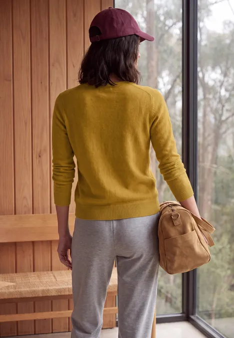 Personne de dos dans une pièce en bois, pull jaune, pantalon gris, casquette bordeaux et un sac beige.
